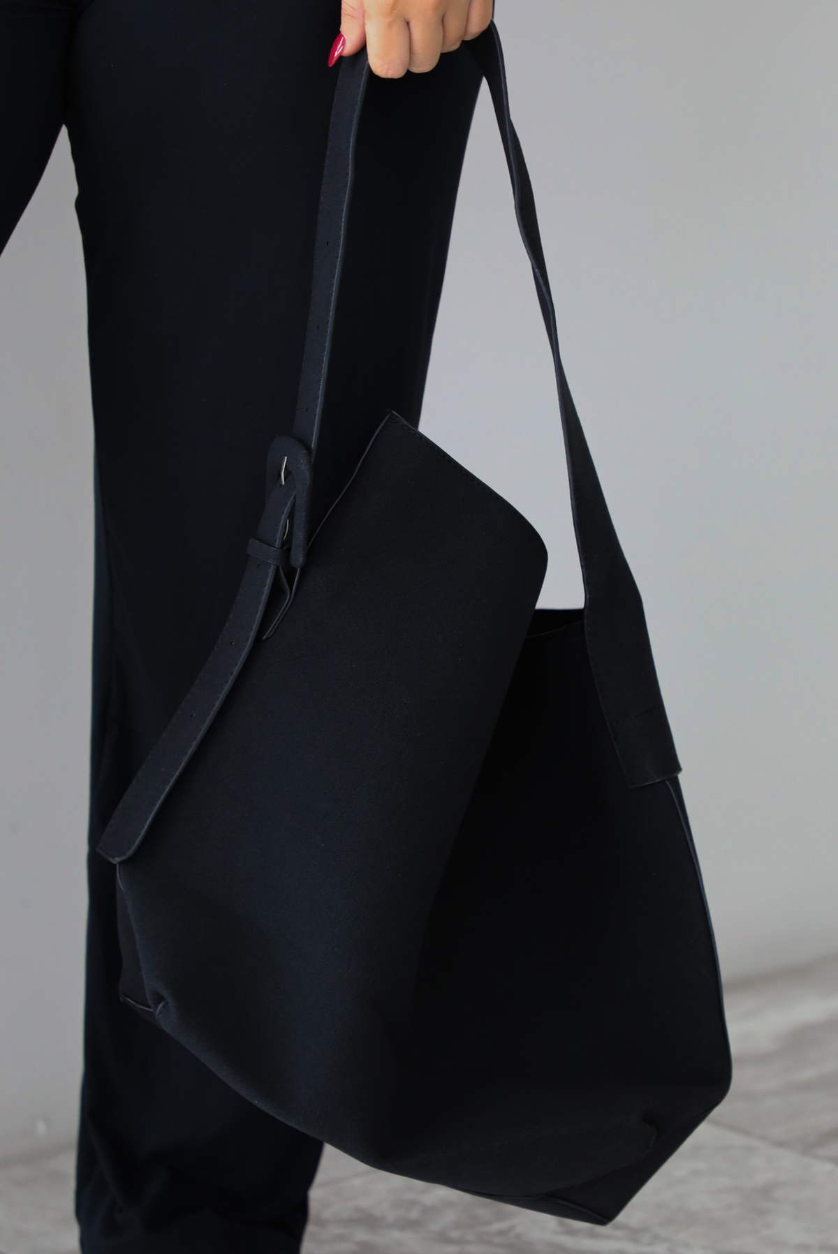 The photo features black shoulder bag that has a thick strap with a buckle closure. The photo is taken against a white wall. 