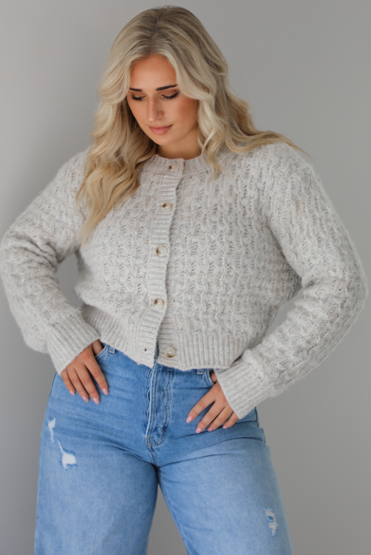 Woman wearing a light grey cardigan buttoned up with high waisted flared hem medium wash denim. The photo shows a closeup of the texture of the cardigan, which resembles a loose textured cable knit. The photo is taken indoors against a white wall.