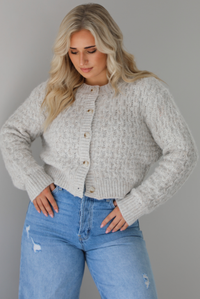Woman wearing a light grey cardigan buttoned up with high waisted flared hem medium wash denim. The photo shows a closeup of the texture of the cardigan, which resembles a loose textured cable knit. The photo is taken indoors against a white wall.