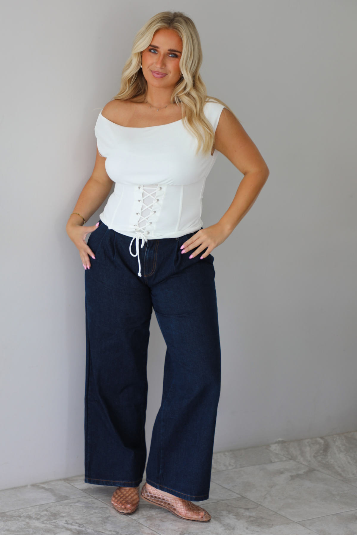A woman modeling a white off-the-shoulder lace-up corset top paired with dark denim wide-leg jeans and pinkish-brown flat shoes, standing indoors against a white wall with white marble flooring. 