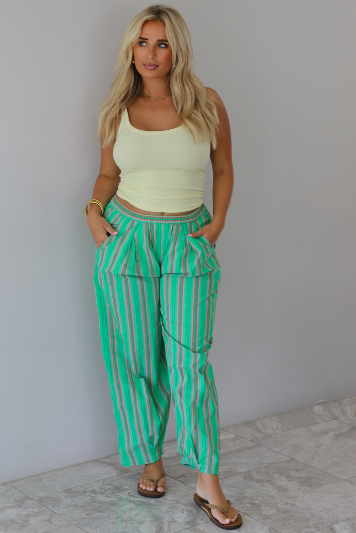 A woman wearing a pale yellow tank and mid-rise pants that have a green, brown, lime green, and tan striped pattern, which have an elastic waistline, functional pockets on either side, and cinched details at the ankles, standing indoors against a white wall with white marble flooring. 