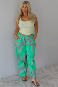 A woman wearing a pale yellow tank and mid-rise pants that have a green, brown, lime green, and tan striped pattern, which have an elastic waistline, functional pockets on either side, and cinched details at the ankles, standing indoors against a white wall with white marble flooring. 