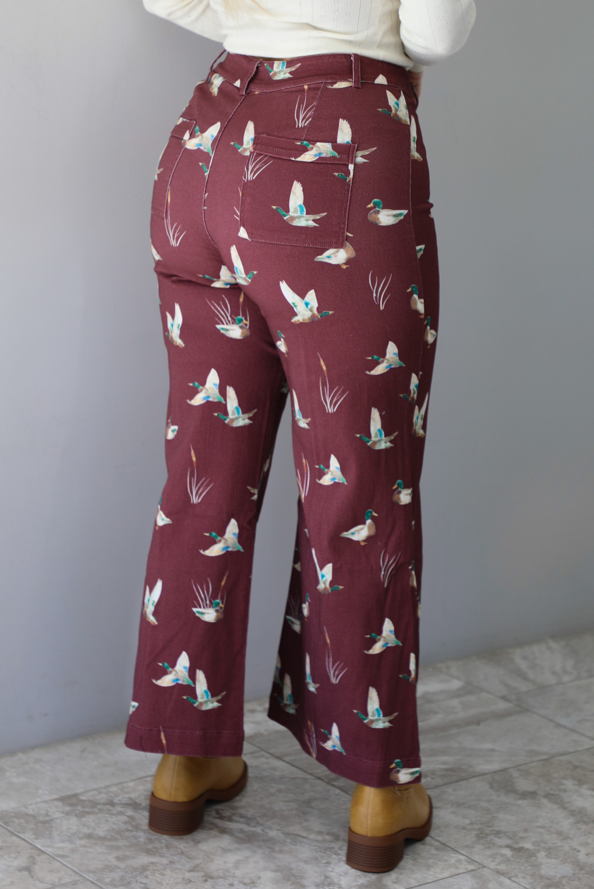 A closeup of a woman from behind wearing a pair of maroon denim pants with a multi-colored repeating mallard design throughout have have a high waisted design, pleated details on the front, standing indoors against a white wall with white marble flooring. 