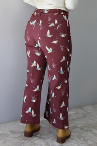 A closeup of a woman from behind wearing a pair of maroon denim pants with a multi-colored repeating mallard design throughout have have a high waisted design, pleated details on the front, standing indoors against a white wall with white marble flooring. 