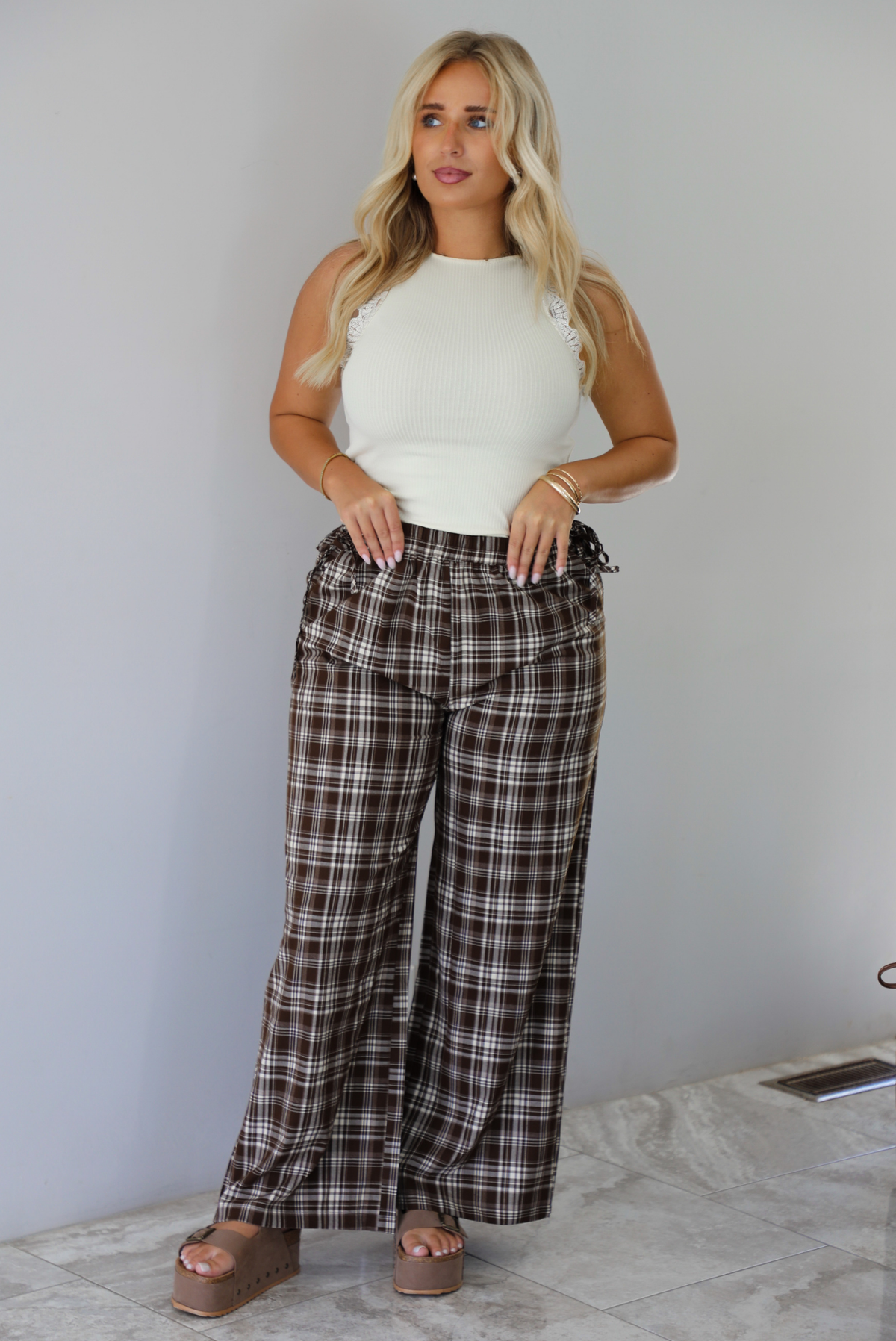 A woman modeling a pair of brown and sand plaid wide-leg pants with a cream ribbed sleeveless top, standing indoors against a white wall with white marble flooring. 