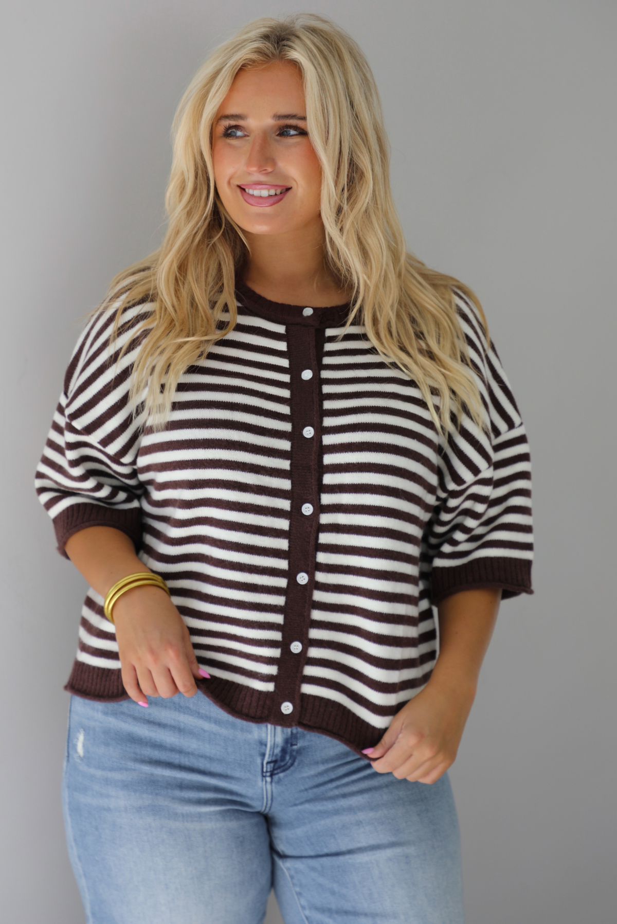 A woman wearing a brown and white striped knit button top cardigan top, paired with light wash denim distressed denim, standing indoors against a white wall.