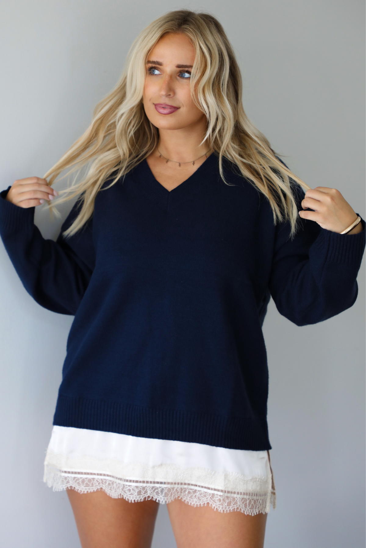 A woman wearing a navy sweater dress with a cream lace trim detail peaking out from underneath the dress giving it a layered look, standing indoors against a white wall.