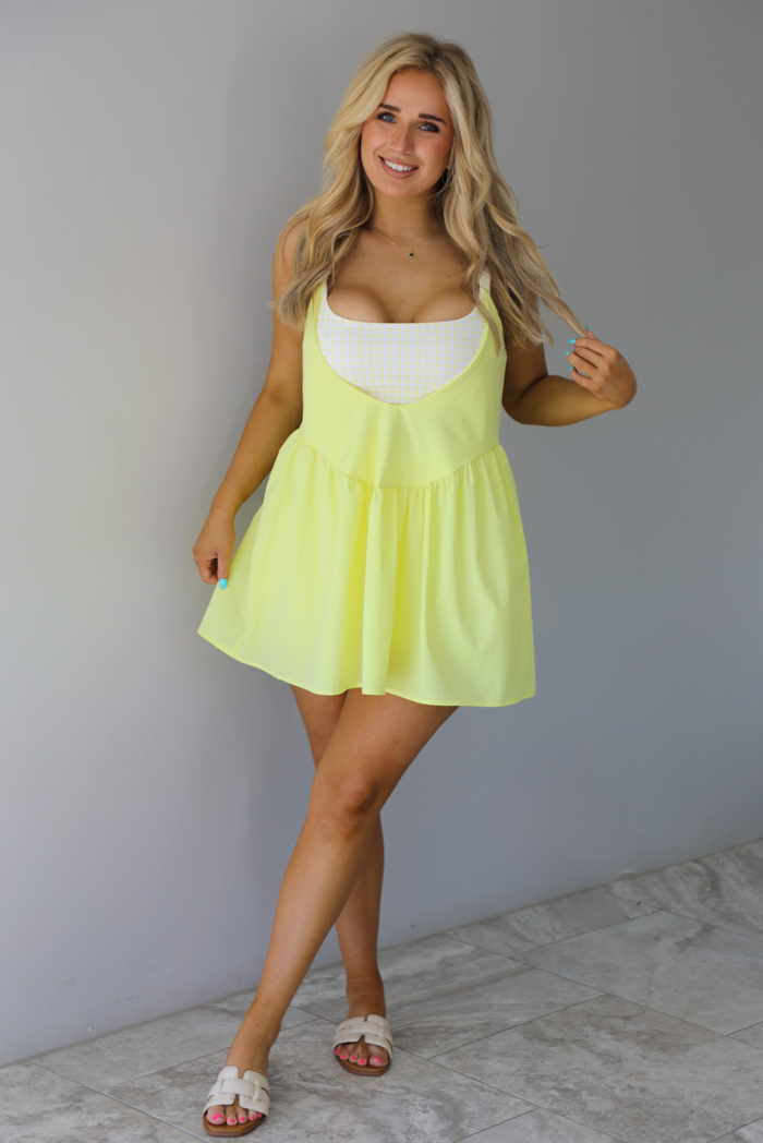 Woman wearing a yellow dress with a yellow and white gingham romper underneath. The photo is taken indoors against a white wall with white marble flooring. 
