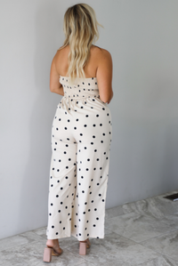 A woman shown from behind wearing a sand strapless jumpsuit with a black polka dot print pattern throughout, standing indoors against a white wall with white marble flooring.
