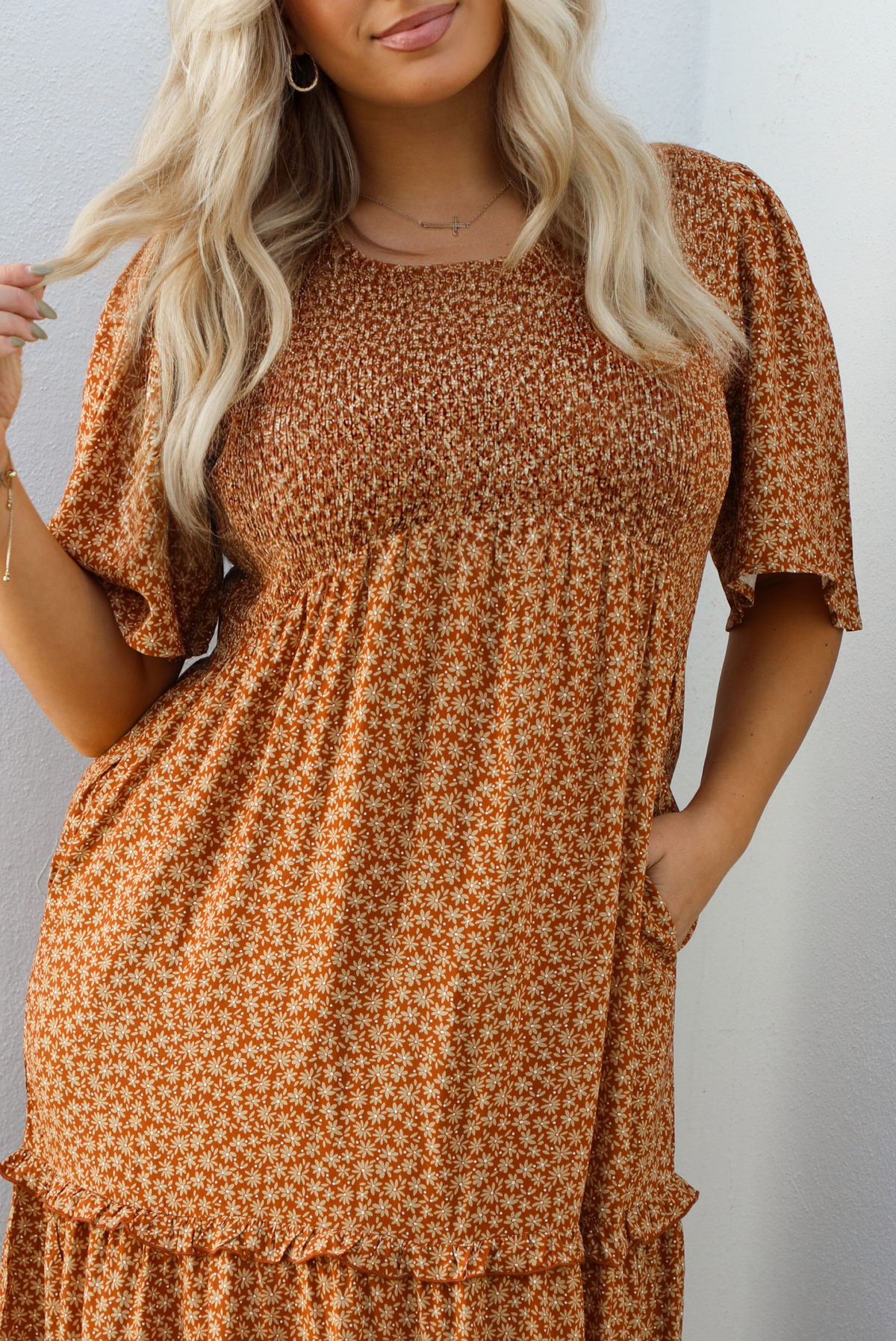 Up close of a woman model outdoors wearing a rust dress with a cream floral print pattern throughout. It has flowy short sleeves, a smocked bodice, and a flowy maxi length skirt with functional pockets on either side. 