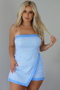 Woman wearing a blue and white strapless striped romper that looks like a dress with a folded over design in the front indoors against a white wall.