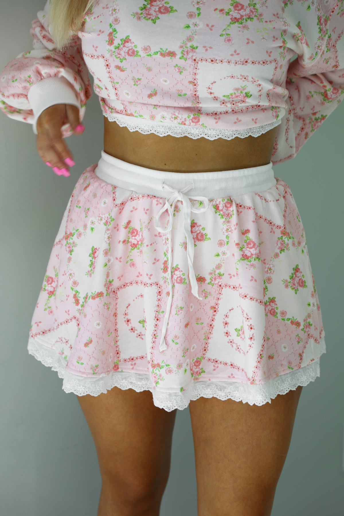 Woman wearing a two piece set featuring a pink floral set that has an oversized long sleeved top and high waisted skirt. The photo shows a closeup of the skirt, which has a drawstring and elastic waistline. The photo is taken indoors against a white wall.  