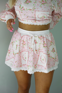 Woman wearing a two piece set featuring a pink floral set that has an oversized long sleeved top and high waisted skirt. The photo shows a closeup of the skirt, which has a drawstring and elastic waistline. The photo is taken indoors against a white wall.  