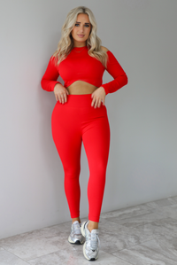 Woman wearing a two piece athletic set that has a cropped top with long sleeves, cold shoulder cut out details, and a cropped cut, with high waisted, fitted leggings. The set is modeled with a pair of silver sneakers. The photo is taken indoors against a white wall with white marbled flooring.