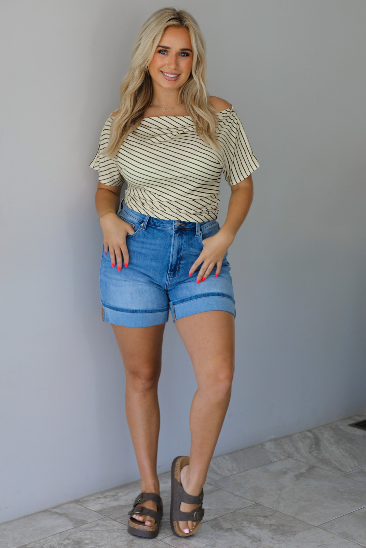 Woman wearing a pale yellow top with a taupe striped pattern with medium wash denim shorts indoors against a white wall with white marble flooring.