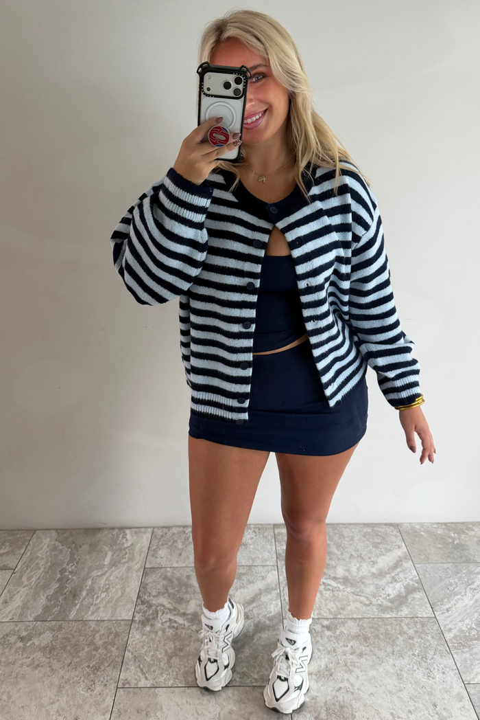 A woman taking a mirror selfie wearing a navy and blue striped cardigan paired with a navy two piece set underneath standing indoors against a white wall with white marble flooring. 