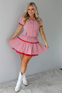 Woman wearing a red and white gingham patterned set that has a short sleeved button-up top that has pleated details at the hem of the top and a high waisted skirt with red lace detailing at the hem. The set is paired with white cowboy boots. The photo is taken indoors against a white wall with white marble flooring. 