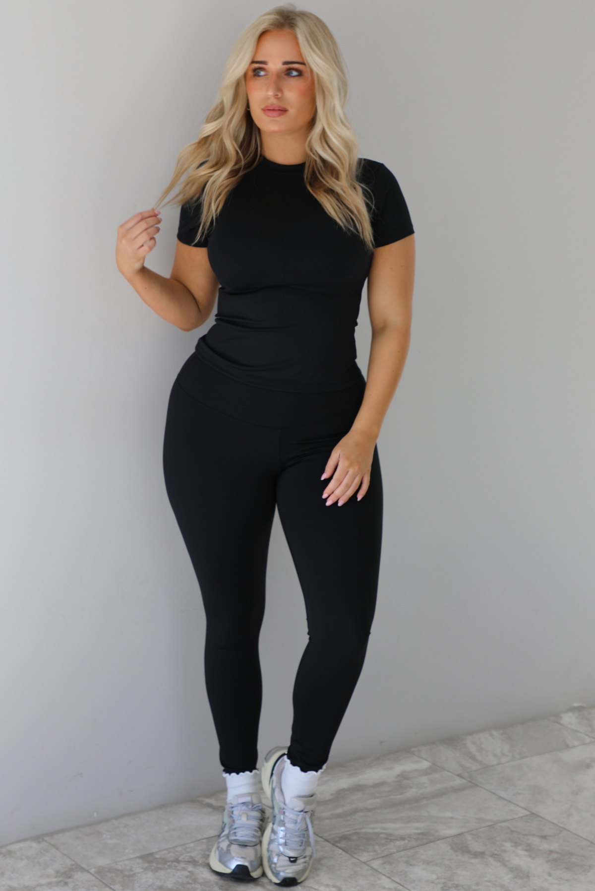 Woman wearing a fitted two piece black set featuring a short sleeved top and fitted leggings. The set is styled with a pair of white ankle socks and silver sneakers. The photo is taken indoors against a white wall with white marble flooring. 
