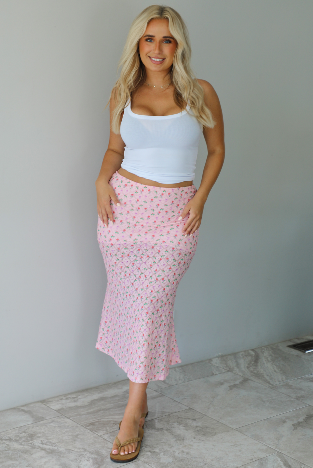 Woman wearing a white tank and pink midi length skirt that has a repeating floral pattern throughout indoors against a white wall with white marble flooring. 