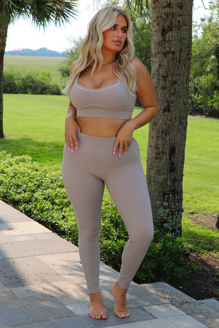 A woman modeling a matching tan activewear set consisting of a scoop-neck sports bra and high-waisted leggings, standing outdoors on a stone walkway with a green lawn and trees in the background.