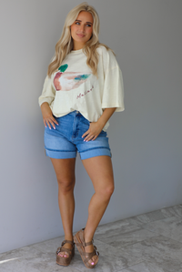 Woman wearing an off-white t-shirt with a graphic design of a mallard duck on the front cuffed into blue denim shorts. The photo is taken indoors against a white wall with white marble flooring. 