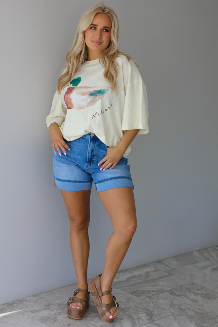 Woman wearing an off-white t-shirt with a graphic design of a mallard duck on the front cuffed into blue denim shorts. The photo is taken indoors against a white wall with white marble flooring. 