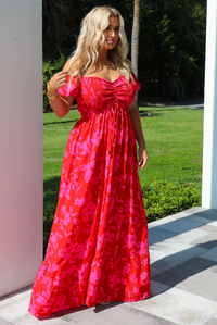 A woman wearing a red and pink floral maxi dress, standing outdoors on a tiled patio with a green lawn and trees in the background.