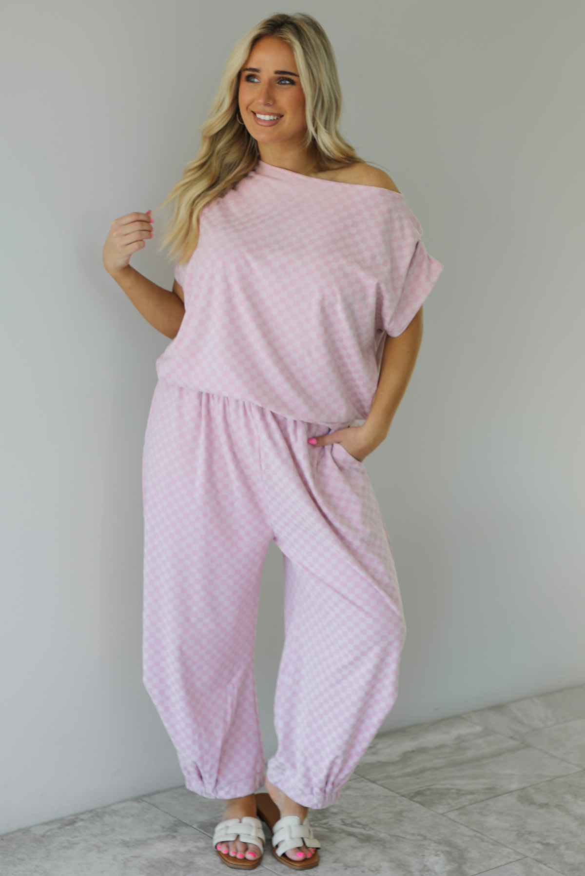 Woman wearing a pink checkered set featuring a slouchy top and pants. The photo is taken indoors against a white wall with white marble flooring.