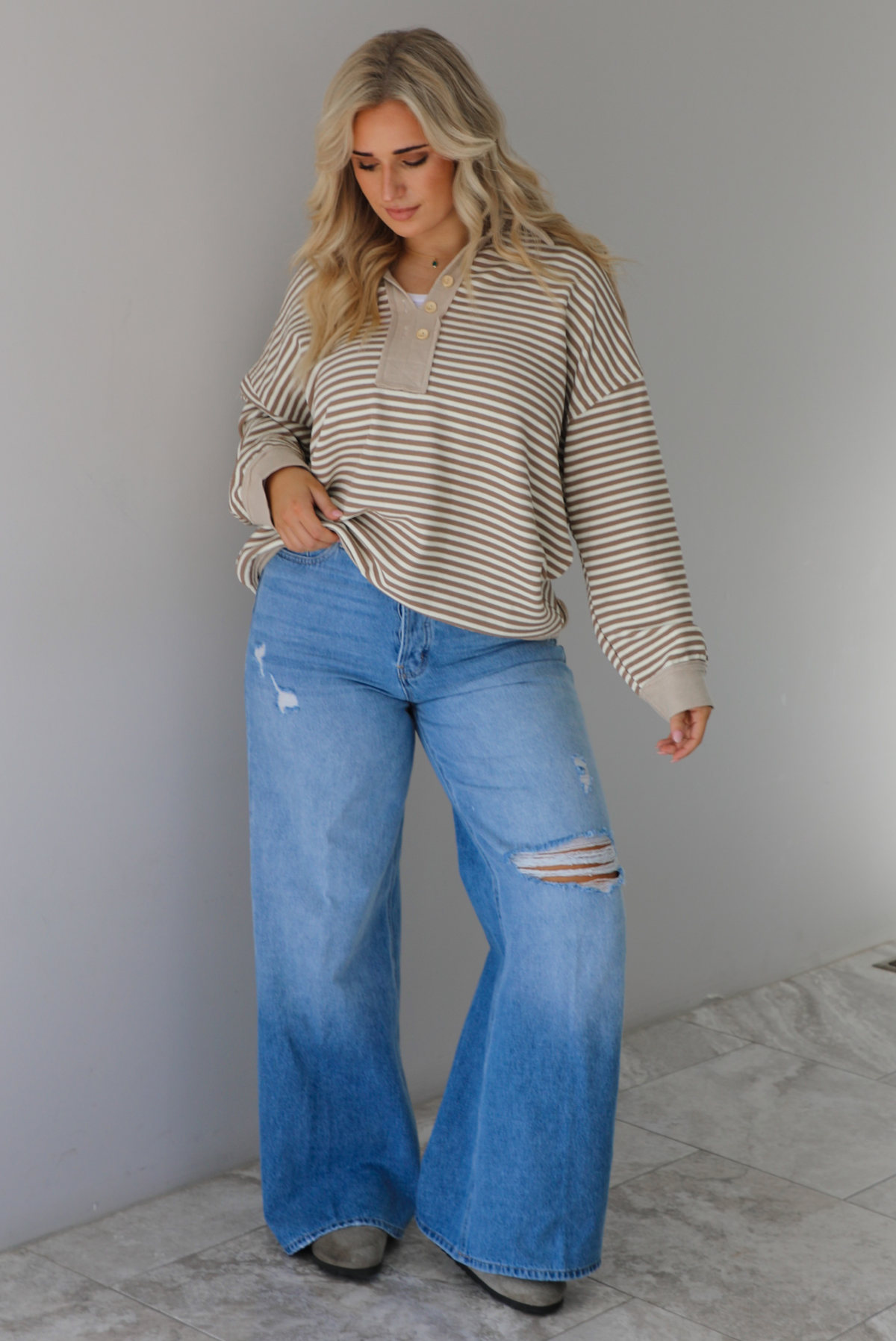Woman wearing a mocha and cream striped pullover with a collared neckline, quarter button closures in the front, that has an oversized fit. The pullover is paired with a medium wash denim. The photo is taken indoors against a white wall with white marble flooring. 