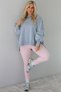 Woman wearing a grey colored oversized sweatshirt with exposed seam details and a high-low design with baby pink leggings. The outfit is styled with white ankle socks and silver sneakers. The photo is taken indoors against a white wall with white marble flooring. 
