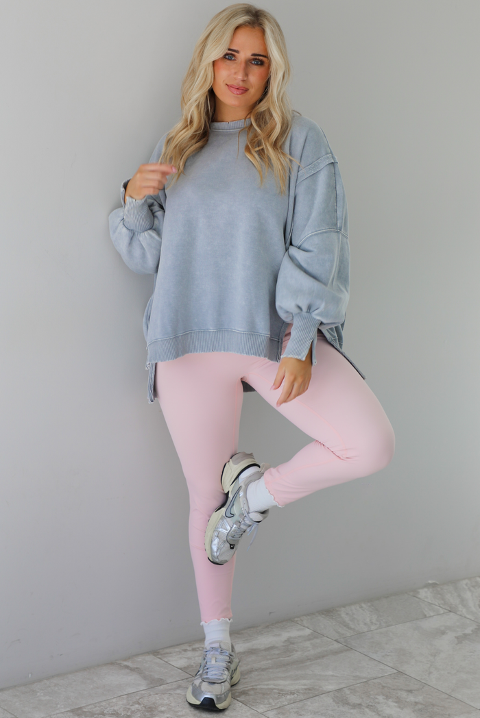 Woman wearing a grey colored oversized sweatshirt with exposed seam details and a high-low design with baby pink leggings. The outfit is styled with white ankle socks and silver sneakers. The photo is taken indoors against a white wall with white marble flooring. 