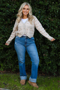 Woman model wearing a grey cardigan that has an orange and white flora print pattern throughout, long sleeves, an asymmetrical hem, and functional button closures in the front, with a long sleeved white top, medium wash denim jeans, and brown shoes in front of a green hedge outdoors. 