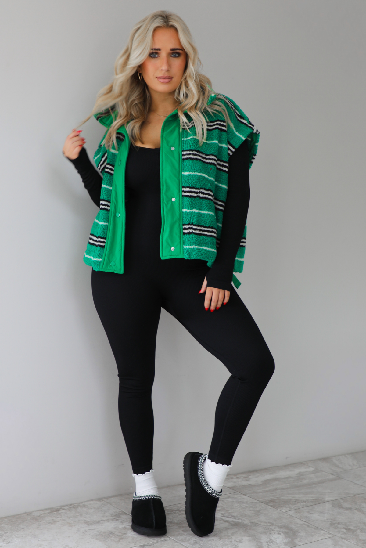 Woman wearing a green sherpa vest with black and white stripes that has an oversized fit, with a long sleeved, fitted, full body jumpsuit, with white slouchy socks and black ugg boot slides. The photo is taken indoors against a white wall with white marbled flooring. 