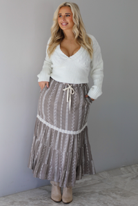 A woman wearing an ivory v-neck sweater paired with a taupe and white embroidered maxi skirt, standing indoors against a white wall with white marble flooring. 