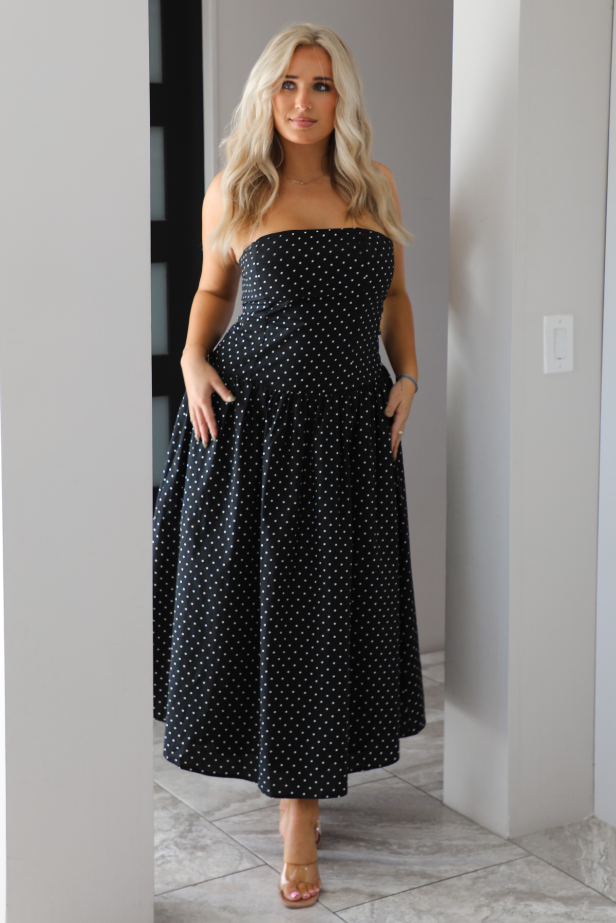 Woman model wearing a strapless/sleeveless, black with white polka dots, maxi length dress indoors. It has a fitted bodice with a flowy skirt. 