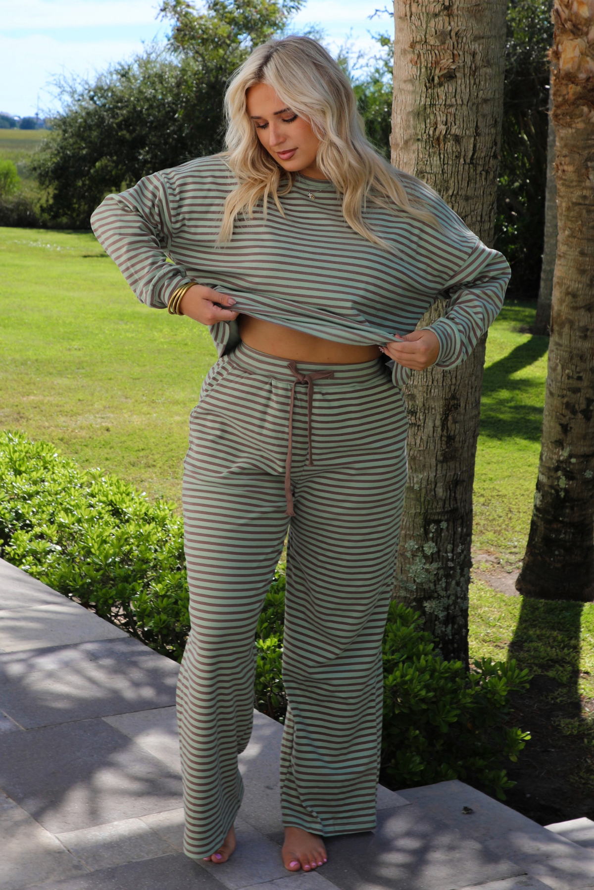 A woman wearing a pale green and brown striped two piece set featuring an oversized long sleeved top and high waisted, wide leg pants, standing outdoors on a stone path with palm trees and other greenery in the background. 