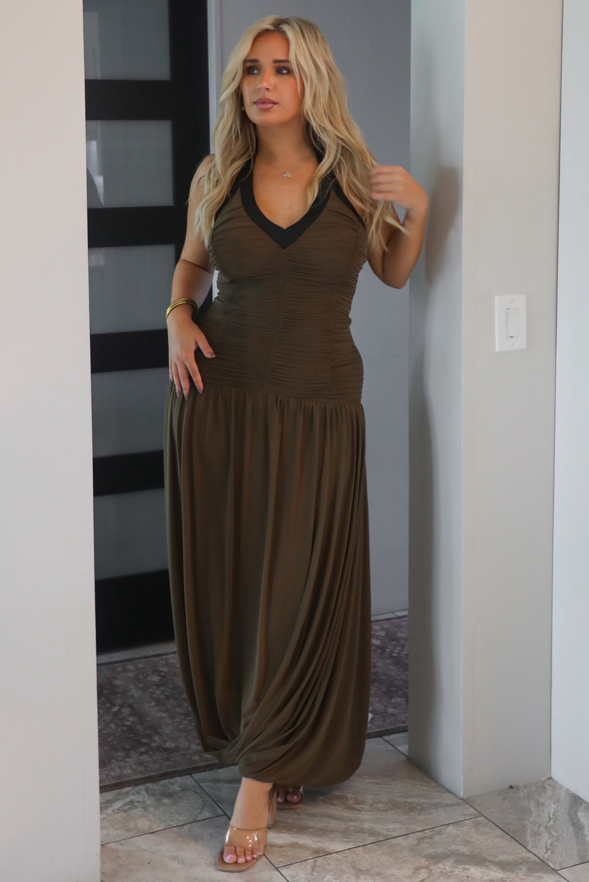 A woman wearing an olive maxi dress with black trim detailing at the v-halter neckline, has a fitted bodice and drop waist design, with a maxi-length flowy skirt, standing in an archway indoors with a black door in the background. 