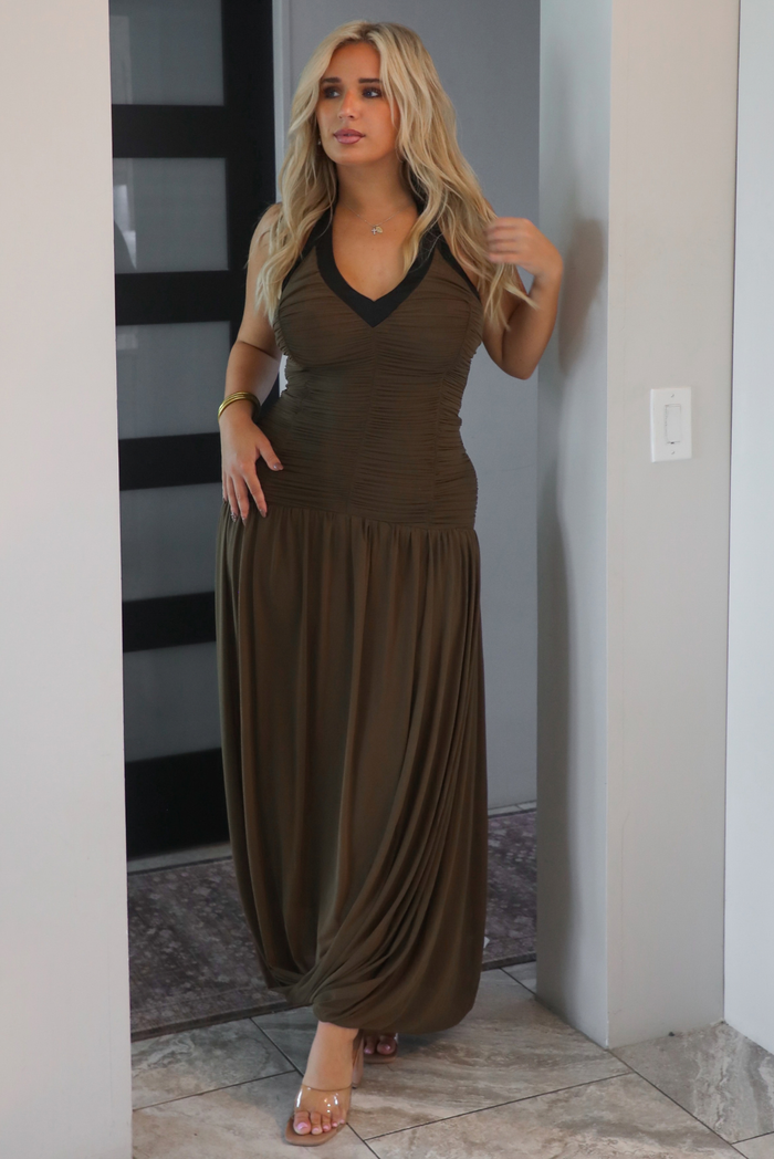 A woman wearing an olive maxi dress with black trim detailing at the v-halter neckline, has a fitted bodice and drop waist design, with a maxi-length flowy skirt, standing in an archway indoors with a black door in the background. 