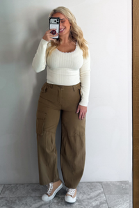 A woman taking a mirror selfie wearing a cream knit long sleeved top and high waisted olive-khaki cargo pants, standing indoors against a white wall with white marble flooring. 