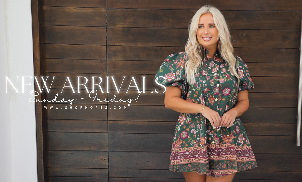 A woman with long blonde hair stands in front of a dark wood-paneled wall wearing a short-sleeve green floral dress with a patterned hem. Large white text on the left reads 'NEW ARRIVALS Sunday - Friday!' with a website URL below.