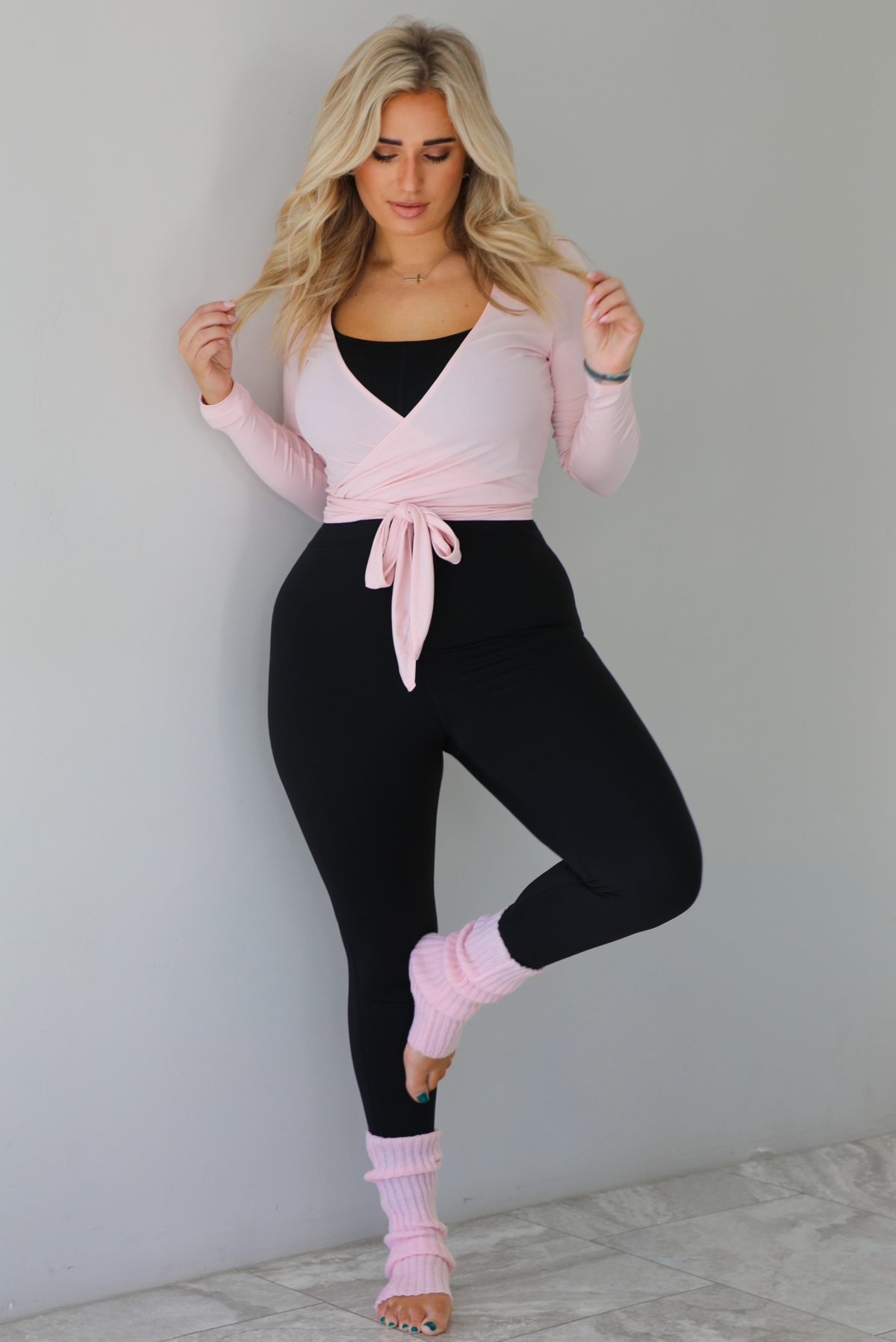 Woman wearing a pink cropped wrap style fitted top over a black fitted jumpsuit. The outfit is paired with pink leg warmers. The photo is taken indoors against a white wall with white marble flooring. 