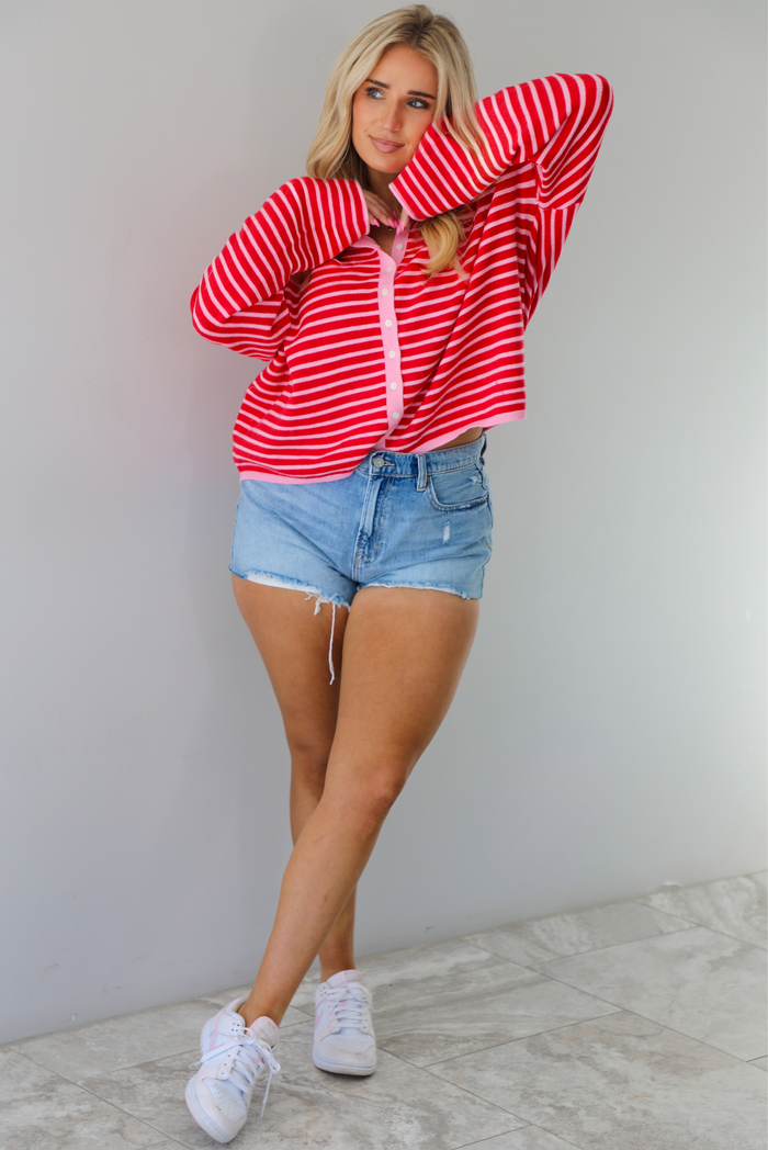 Woman wearing a red and pink striped oversized cardigan with light wash denim shorts. The photo is taken indoors against a white wall with white marble flooring. 