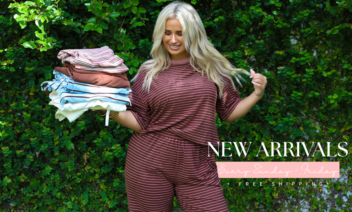 A woman standing in front of a green plant covered hedge wearing a brown and pink two piece set holding other sets folded up in her hand. Promotional text in the bottom right corner reads "NEW ARRIVALS EVERY SUNDAY - FRIDAY + FREE SHIPPING".