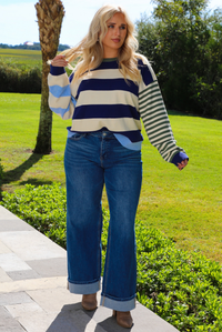 Woman wearing a navy, cream, blue, and green alternating striped design throughout, paired with dark denim jeans, standing outdoors on a stone path with palm trees and other greenery in the background. 