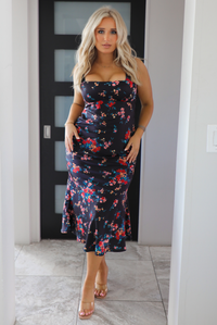 Woman model wearing a navy dress with a multi-colored floral print. The dress is a satin material, is a midi length, and is curve-hugging. The photo is taken inside a modern white and black interior. 