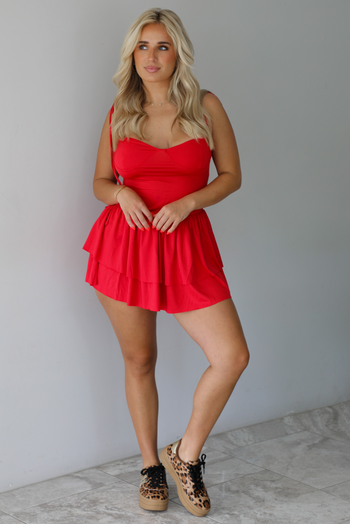 Woman wearing a red mini dress that has thin, spaghetti straps, a fitted bodice, and a flowy, ruffled skirt indoors against a white wall with white marble flooring. 