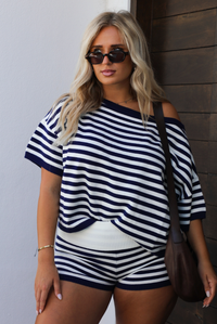 A woman wearing a navy and white horizontally striped off-the-shoulder top and matching striped shorts, accessorized with a brown shoulder bag and sunglasses.