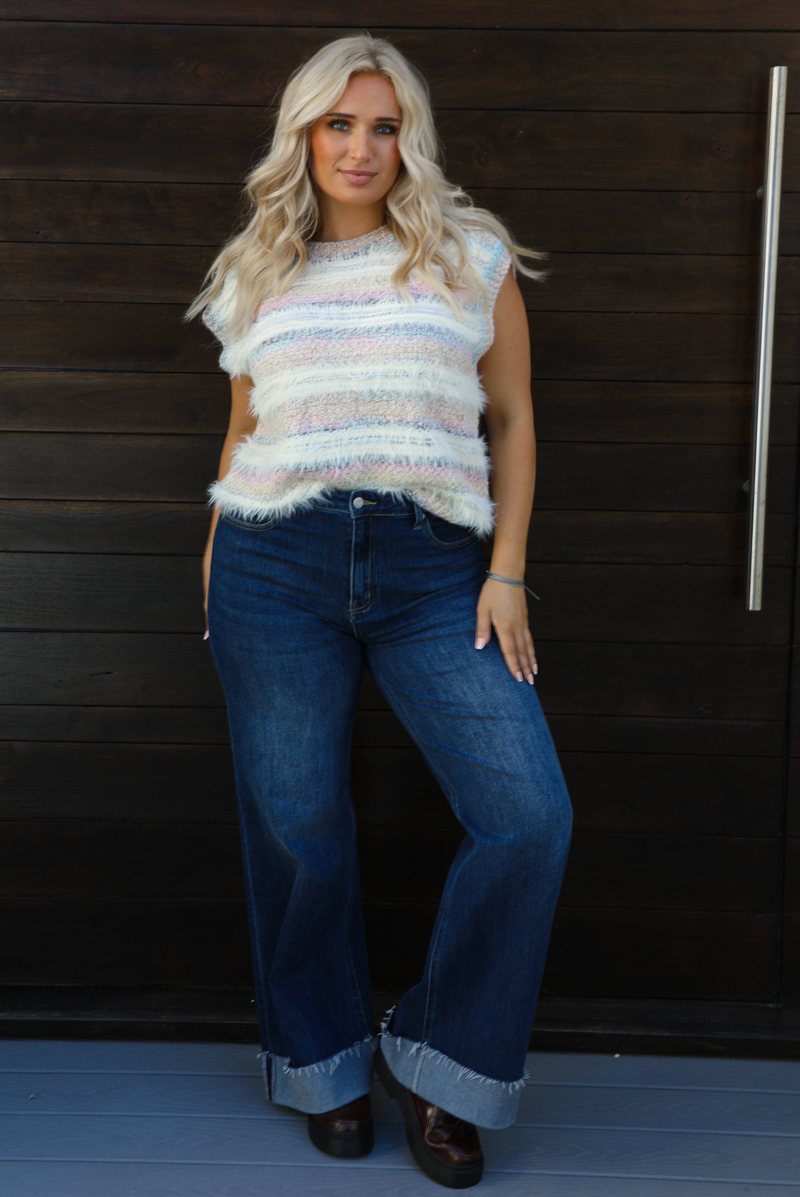 Woman model wearing a soft, striped sweater vest with dark wash denim jeans. The photo is taken outdoors against a dark brown wooden door. 