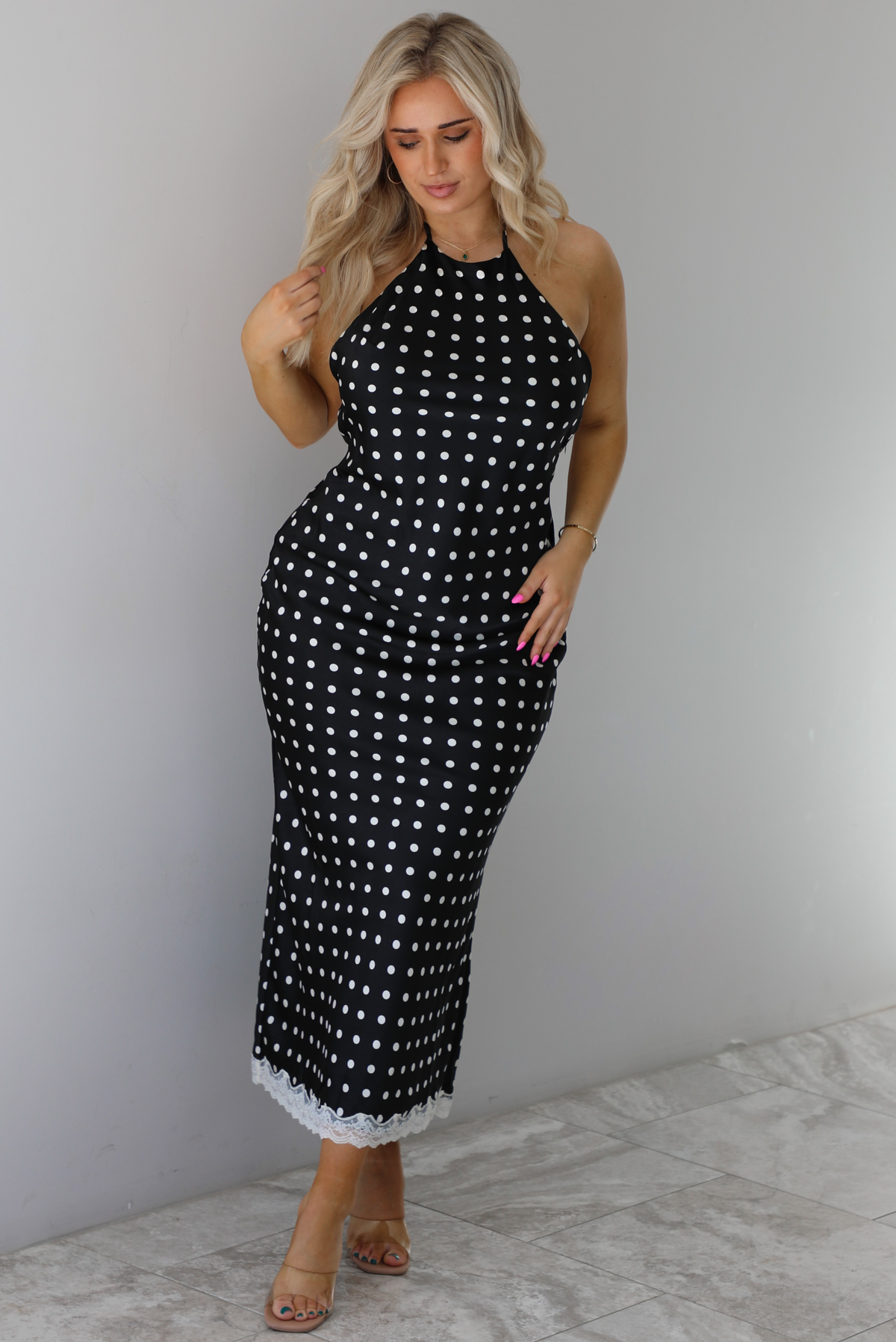 Woman wearing a black wearing a white polka dot printed midi dress that has a halter tie neckline, is form fitting, with white lace trim at the bottom. The photo is taken indoors against a white wall with white marble flooring. 