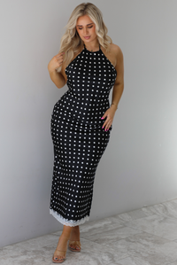 Woman wearing a black wearing a white polka dot printed midi dress that has a halter tie neckline, is form fitting, with white lace trim at the bottom. The photo is taken indoors against a white wall with white marble flooring. 